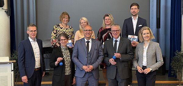 9 Personen auf einer Bühne in einer Schulaula